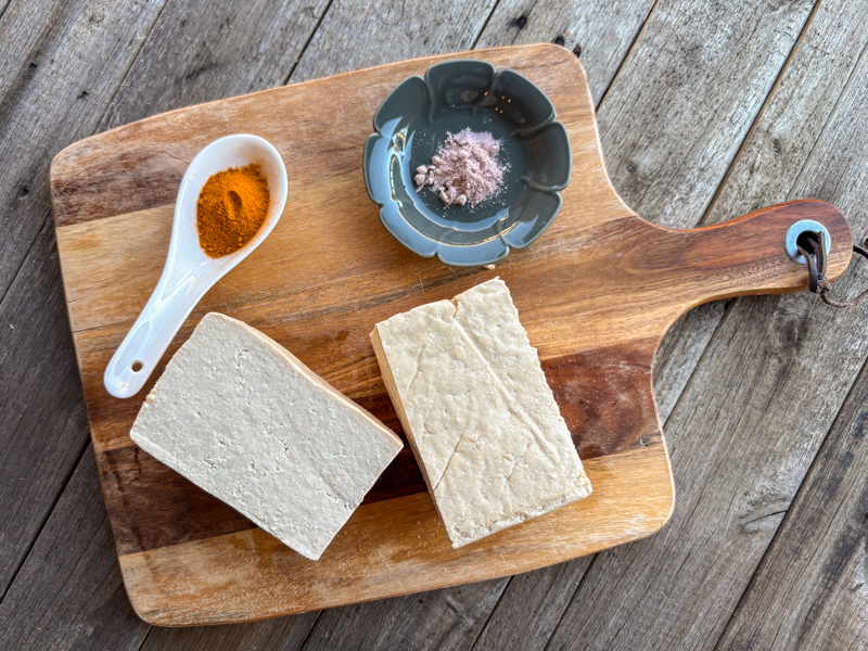 Image of wooden cutting board with hard tofu, as well as black salt and turmeric displayed nicely.